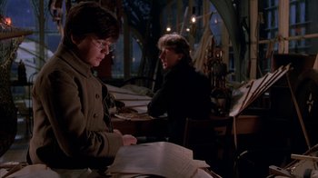Movie still from “Young Sherlock Holmes” (1985), directed by Barry Levinson – A man sitting at a table looking at papers with another man standing next to him; Medium shot, Over the shoulder angle