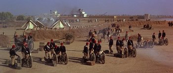 Movie still from “Young Winston” (1972), directed by Richard Attenborough – A group of men sitting next to each other on top of a dirt field; Extreme Wide shot, High angle