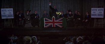Movie still from “Young Winston” (1972), directed by Richard Attenborough – A group of people standing around a stage with a british flag on it; Wide shot, Low angle