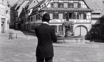 Movie still from “Yoyo” (1965), directed by Pierre Étaix – A man standing in front of a fountain in front of a building; Wide shot, High angle