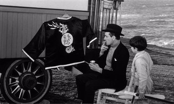 Movie still from “Yoyo” (1965), directed by Pierre Étaix – A man sitting in front of a jacket on a rack; Medium shot, Over the shoulder angle