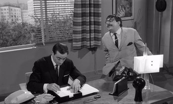 Movie still from “Yoyo” (1965), directed by Pierre Étaix – A man sitting at a desk writing on a piece of paper next to another man; Medium shot, Over the shoulder angle