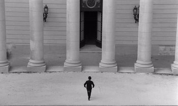 Movie still from “Yoyo” (1965), directed by Pierre Étaix – A man in a black suit walking in front of a building with columns; Extreme Wide shot, High angle