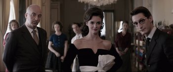 Movie still from “Yves Saint Laurent” (2014), directed by Jalil Lespert – A woman in a black dress standing in front of a group of people; Medium shot, Over the shoulder angle