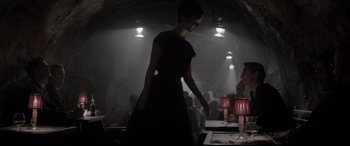 Movie still from “Yves Saint Laurent” (2014), directed by Jalil Lespert – A woman in a black dress standing next to a table; Medium shot, Over the shoulder angle