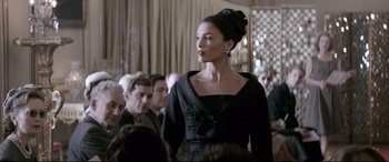 Movie still from “Yves Saint Laurent” (2014), directed by Jalil Lespert – A woman in a black dress standing in front of a group of people; Medium shot, Over the shoulder angle