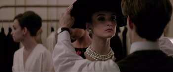 Movie still from “Yves Saint Laurent” (2014), directed by Jalil Lespert – A woman in a hat and pearls holding her hat; Close Up shot, Over the shoulder angle