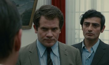 Movie still from “Z” (1969), directed by Costa-Gavras – A man in a suit standing next to another man in a suit; Close Up shot, Over the shoulder angle