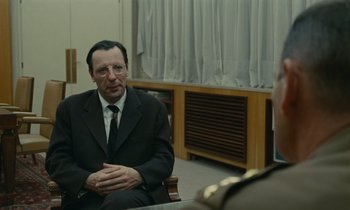 Movie still from “Z” (1969), directed by Costa-Gavras – A man wearing a suit and tie sitting in a chair; Medium shot, Over the shoulder angle