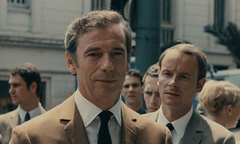 Movie still from “Z” (1969), directed by Costa-Gavras – A man in a suit and tie standing next to another man in a suit and tie; Close Up shot, Over the shoulder angle