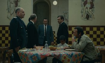 Movie still from “Z” (1969), directed by Costa-Gavras – A group of men standing around a table with food on it; Wide shot, High angle