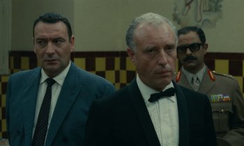 Movie still from “Z” (1969), directed by Costa-Gavras – A man in a suit and tie standing next to another man in a suit and tie; Medium shot, Low angle