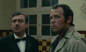 Movie still from “Z” (1969), directed by Costa-Gavras – Two men standing next to each other in front of a building; Medium shot, Low angle