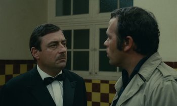 Movie still from “Z” (1969), directed by Costa-Gavras – Two men in suits talking to each other in front of a checkerboard wall; Medium shot, Over the shoulder angle