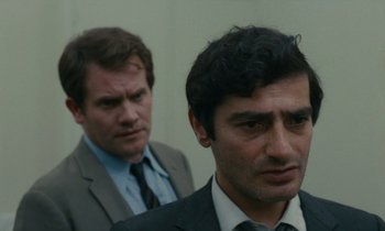 Movie still from “Z” (1969), directed by Costa-Gavras – A man in a suit and tie standing next to another man in a suit and tie; Close Up shot, Over the shoulder angle