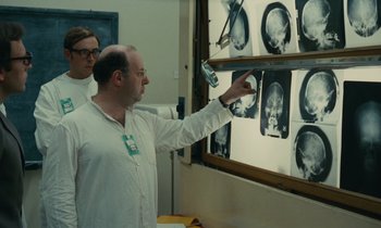 Movie still from “Z” (1969), directed by Costa-Gavras – A man in a white shirt is looking at a picture of a skull; Medium shot, Over the shoulder angle