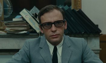 Movie still from “Z” (1969), directed by Costa-Gavras – A man wearing a suit and tie sitting in front of a stack of books; Close Up shot, Low angle