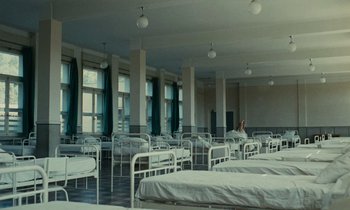 Movie still from “Z” (1969), directed by Costa-Gavras – A room filled with beds and a woman sitting in the middle; Wide shot, Low angle