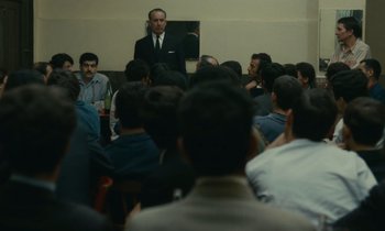 Movie still from “Z” (1969), directed by Costa-Gavras – A group of people sitting in a room with a man in a suit; Wide shot, Over the shoulder angle