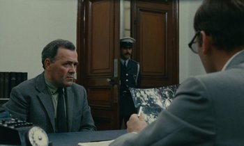 Movie still from “Z” (1969), directed by Costa-Gavras – A man sitting at a table in front of two other men; Medium shot, Over the shoulder angle