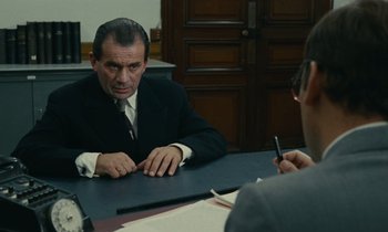 Movie still from “Z” (1969), directed by Costa-Gavras – A man sitting at a table in front of another man holding a pen; Medium shot, Over the shoulder angle