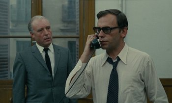 Movie still from “Z” (1969), directed by Costa-Gavras – A man talking on a phone next to another man; Medium shot, Low angle