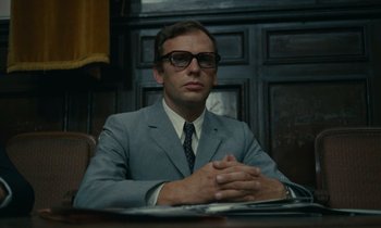 Movie still from “Z” (1969), directed by Costa-Gavras – A man sitting at a table wearing a suit and glasses; Medium shot, Low angle
