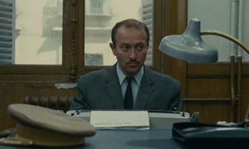 Movie still from “Z” (1969), directed by Costa-Gavras – A man sitting at a typewriter in a suit and tie; Medium shot, Low angle