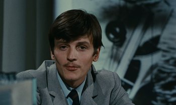 Movie still from “Z” (1969), directed by Costa-Gavras – A man in a suit and tie sitting in front of a picture; Close Up shot, High angle