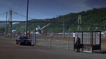 Movie still from “Zero Effect” (1998), directed by Jake Kasdan – A man standing next to a bus stop near a fence; Extreme Wide shot, High angle