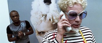 An older woman talking on a phone with a large picture of a woman's face behind her