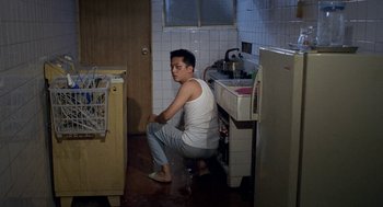 Movie still from “Rebels of the Neon God” (1992), directed by Tsai Ming-liang – A man sitting on the floor of a kitchen next to a sink; Wide shot, High angle
