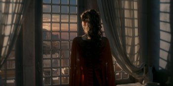 Movie still from “House of the Dragon” (2022), created by George R.R. Martin – A woman standing in front of a window looking out at the sky; Wide shot, Over the shoulder angle