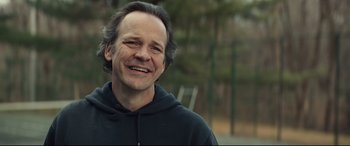 Movie still from “Human Capital” (2019), directed by Marc Meyers – A man with long gray hair smiles for the camera; Close Up shot, Over the shoulder angle
