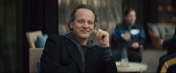 Movie still from “Human Capital” (2019), directed by Marc Meyers – A man in a black jacket is posing for a picture; Close Up shot, Over the shoulder angle