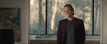 Movie still from “Human Capital” (2019), directed by Marc Meyers – A man standing in front of a window with trees in the background; Medium shot, Low angle
