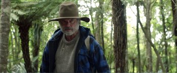 Movie still from “Hunt for the Wilderpeople” (2016), directed by Taika Waititi – An older man with a beard and a hat in the woods; Medium shot, Over the shoulder angle