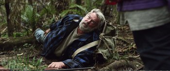 Movie still from “Hunt for the Wilderpeople” (2016), directed by Taika Waititi – An old man with a beard and a backpack laying on the ground in the woods; Medium shot, Over the shoulder angle