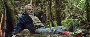 Movie still from “Hunt for the Wilderpeople” (2016), directed by Taika Waititi – An older man sitting on the ground in the woods; Wide shot, Low angle