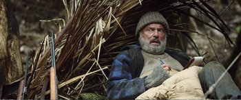 Movie still from “Hunt for the Wilderpeople” (2016), directed by Taika Waititi – A man with a beard and a hat is sitting in a straw hut; Medium shot, Low angle