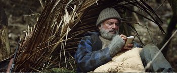 Movie still from “Hunt for the Wilderpeople” (2016), directed by Taika Waititi – A man with a beard and a knitted hat is sitting in front of a straw hut; Medium shot, Low angle