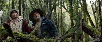 Movie still from “Hunt for the Wilderpeople” (2016), directed by Taika Waititi – An older man in a hat and jacket standing in the woods; Medium shot, Low angle