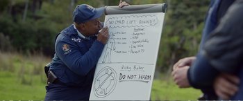 Movie still from “Hunt for the Wilderpeople” (2016), directed by Taika Waititi – A man in a blue jacket is writing on a poster board; Wide shot, High angle