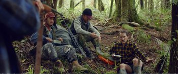 Movie still from “Hunt for the Wilderpeople” (2016), directed by Taika Waititi – A group of men sitting on the ground in the woods; Medium shot, Over the shoulder angle