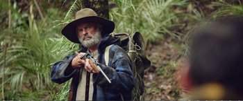 Movie still from “Hunt for the Wilderpeople” (2016), directed by Taika Waititi – An older man with a beard and a hat holding a rifle and wearing a backpack; Medium shot, Over the shoulder angle