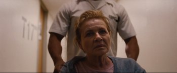 Movie still from “I Care a Lot” (2020), directed by J Blakeson – An older woman looking at the camera with a man standing behind her; Close Up shot, High angle