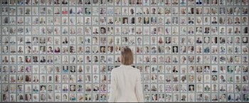 Movie still from “I Care a Lot” (2020), directed by J Blakeson – A woman standing in front of a wall with many pictures of people; Wide shot, High angle