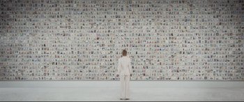 Movie still from “I Care a Lot” (2020), directed by J Blakeson – A woman in a white suit standing in front of a wall of pictures; Extreme Wide shot, High angle
