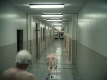 Movie still from “I'm Thinking of Ending Things” (2020), directed by Charlie Kaufman – A man walking down a hall way with a woman in the background; Wide shot, High angle