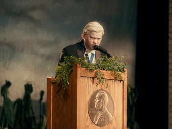 Movie still from “I'm Thinking of Ending Things” (2020), directed by Charlie Kaufman – A man standing at a podium speaking to an audience; Medium shot, High angle